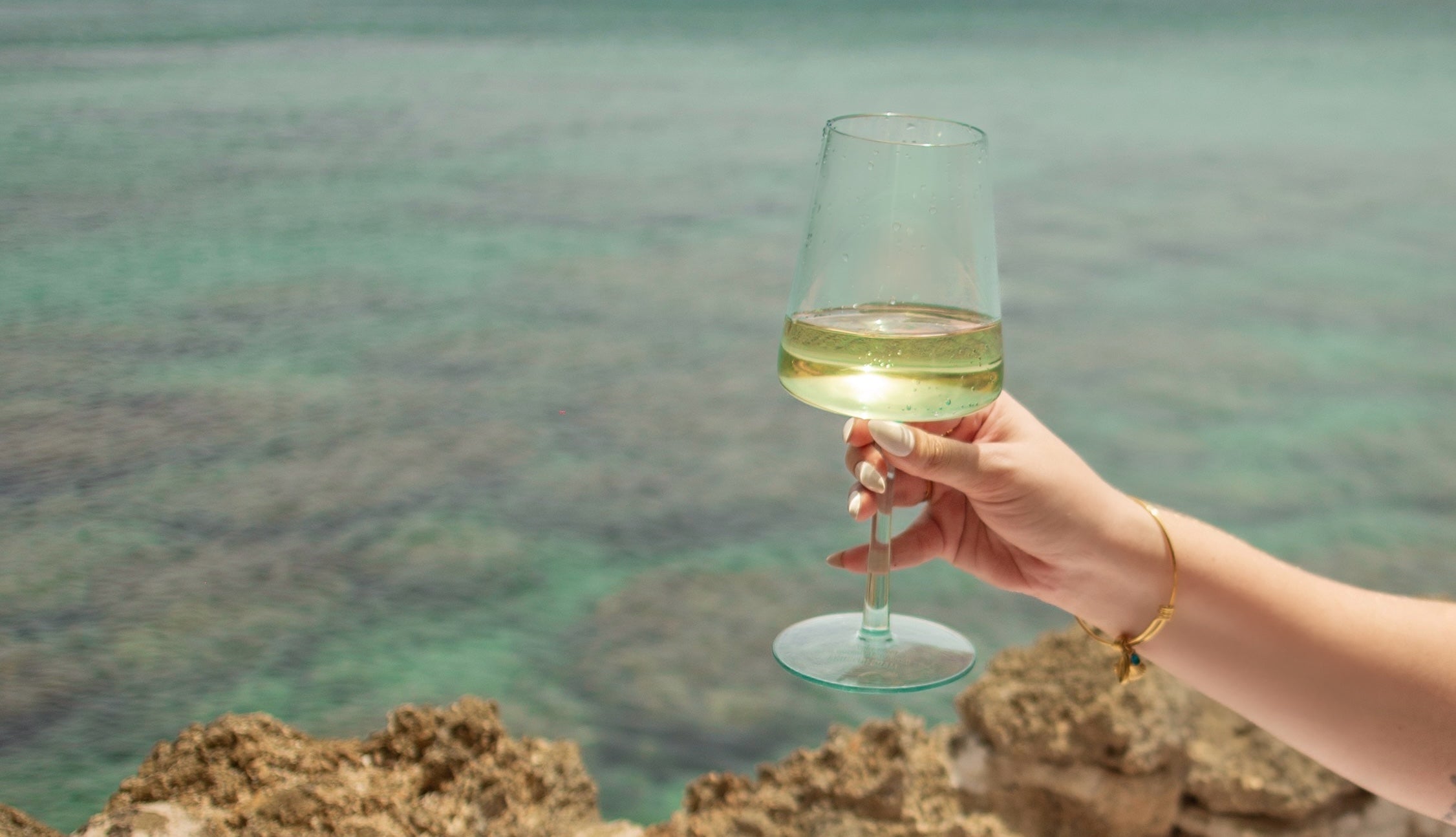 Hand holding a glass of chilled Medly white wine with clear turquoise sea in the background.