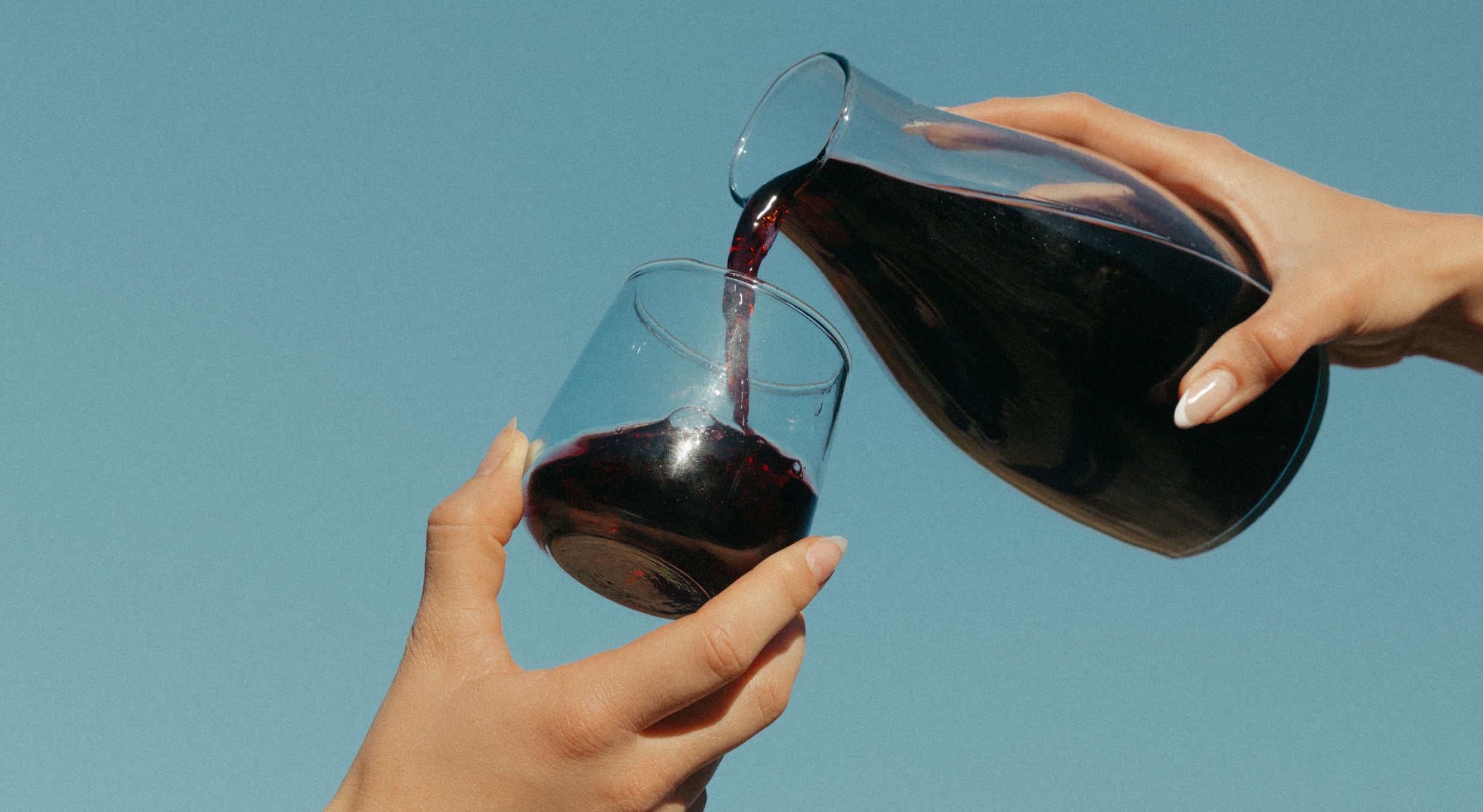  Hands pour Medly red wine from a clear carafe into a stemless glass against a pale blue sky.