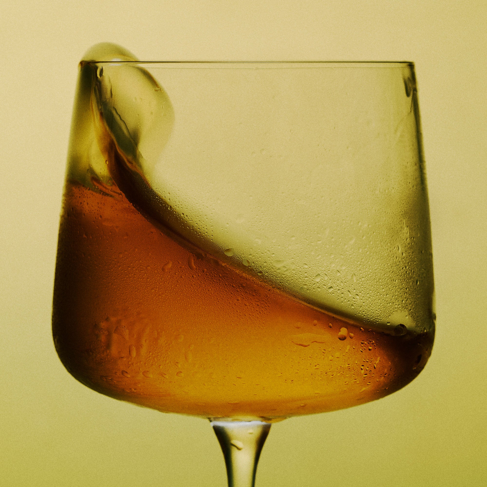 A close-up of a clear stemmed glass containing a Medly orange wine, with condensation droplets on the glass and a soft yellow-green gradient background.